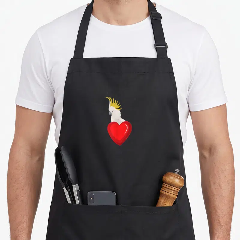 Tablier de cuisine noir avec un perroquet blanc à crête jaune tenant un grand cœur rouge brillant au centre.