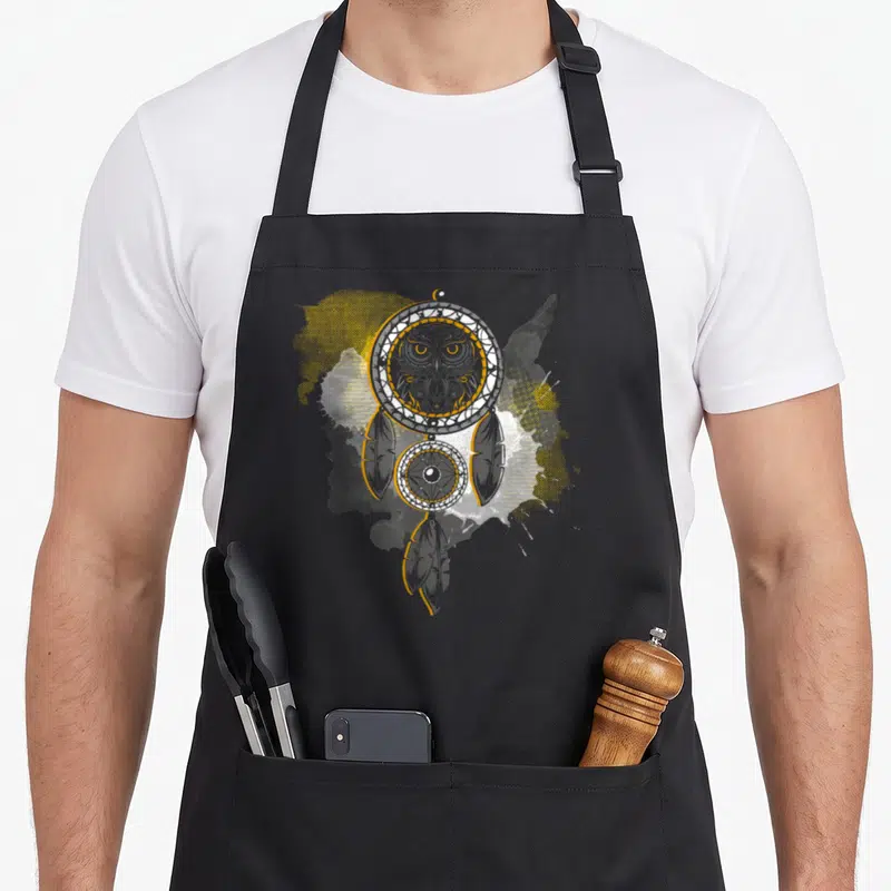Tablier de cuisine noir avec un attrape-rêves stylisé représentant un hibou entouré de plumes et de taches artistiques.