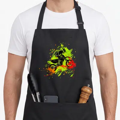 Tablier de cuisine noir avec illustration dynamique d'un pilote de motocross dirt bike sur fond d'éclaboussures vertes et oranges.