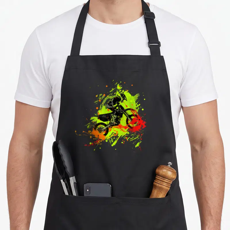 Tablier de cuisine noir avec illustration dynamique d'un pilote de motocross dirt bike sur fond d'éclaboussures vertes et oranges.