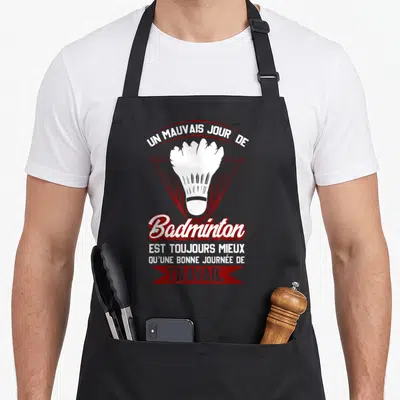 Tablier de cuisine noir avec un volant de badminton blanc encadré par des textes humoristiques en rouge et blanc.