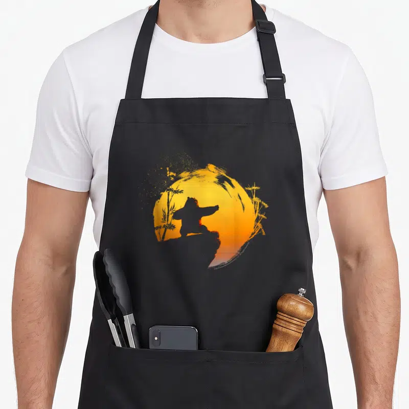Tablier de cuisine noir avec silhouette d'ours pratiquant le Tai Chi devant un soleil orange et des tiges de bambou dorées.