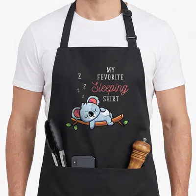 Tablier de cuisine noir avec illustration d'un koala gris dormant sur une branche et du texte en anglais My Favorite Sleeping Shirt.