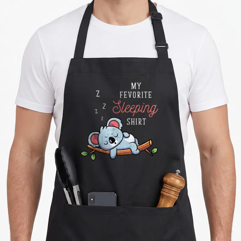Tablier de cuisine noir avec illustration d'un koala gris dormant sur une branche et du texte en anglais My Favorite Sleeping Shirt.