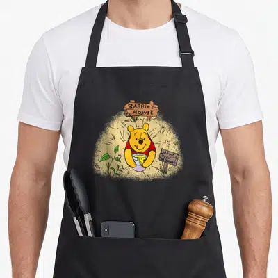Tablier de cuisine noir avec Winnie l'ourson tenant un pot de miel et deux panneaux en bois rustiques.
