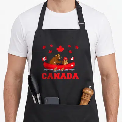 Tablier de cuisine noir avec ours et castor en canoë rouge, feuilles d'érable et texte CANADA en lettres capitales rouges.