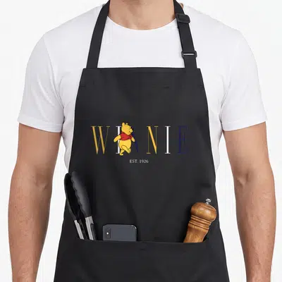 Tablier de cuisine noir avec Winnie l'ourson traversant le texte Est 1926 en lettres colorées