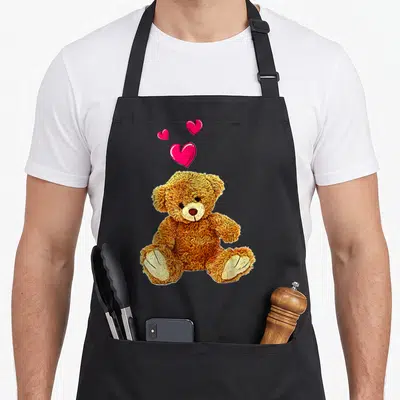 Tablier de cuisine noir avec ours en peluche dorée et trois cœurs roses flottant au-dessus.