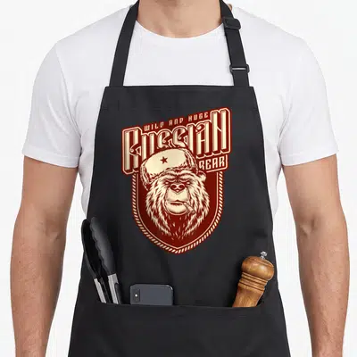 Tablier de cuisine noir avec illustration d'un ours russe vintage coiffé d'une Ushanka et fumant un cigare dans un style badge rouge et crème.