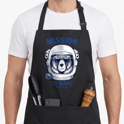 Tablier de cuisine noir avec un ours astronaute en lunettes de soleil et le texte Mission to Mars Super Dude imprimé en bleu.