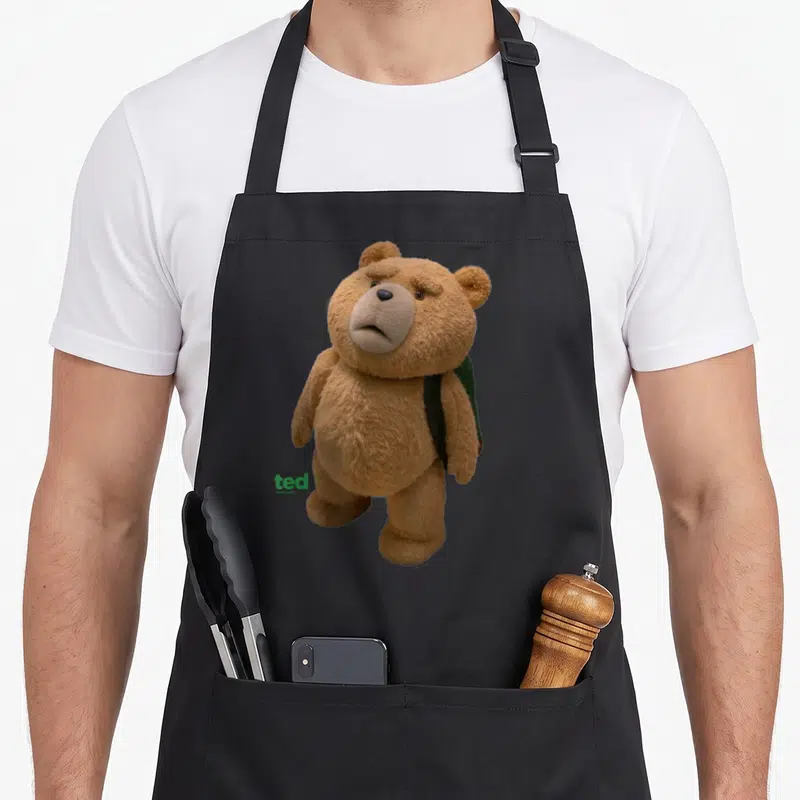 Tablier de cuisine noir avec ours en peluche Ted portant un sac à dos vert et logo vert en bas à gauche.