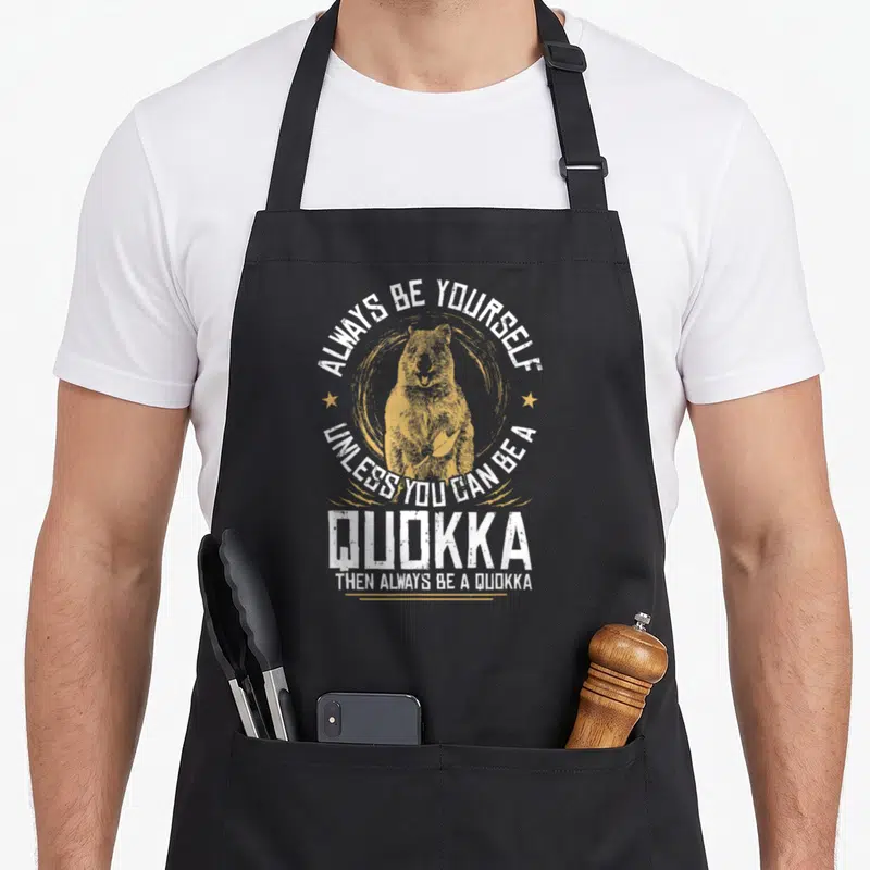 Tablier de cuisine noir avec illustration jaune d'un quokka souriant entouré de texte blanc humoristique.
