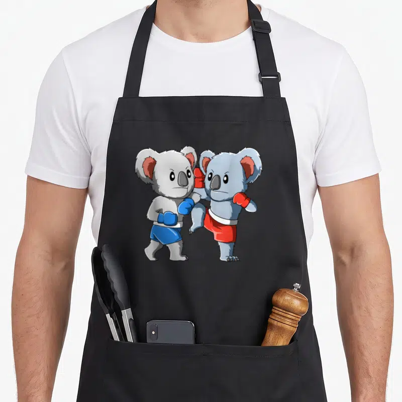 Tablier de cuisine noir avec illustration de deux koalas s'affrontant en boxe thaïlandaise, gants et shorts colorés.