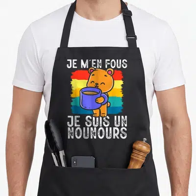 Tablier de cuisine noir avec ours orange tenant une tasse bleue sur fond arc-en-ciel et texte blanc.