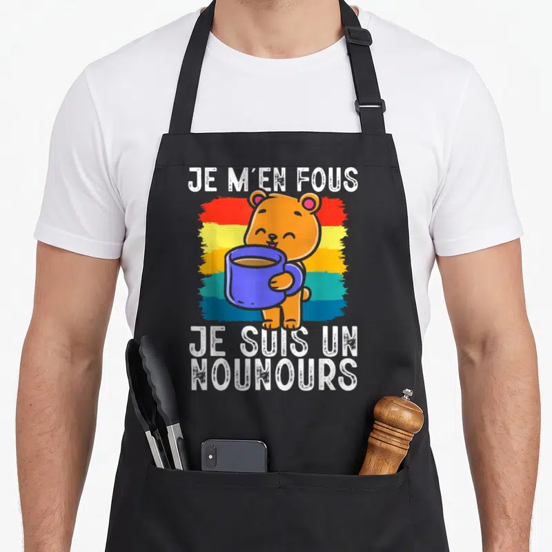 Tablier de cuisine noir avec ours orange tenant une tasse bleue sur fond arc-en-ciel et texte blanc.