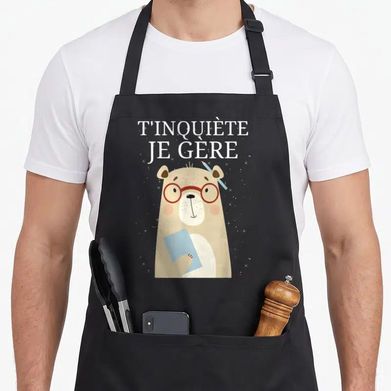 Tablier de cuisine noir avec ours beige à lunettes rouges tenant une feuille bleue et le texte T'inquiète Je Gère