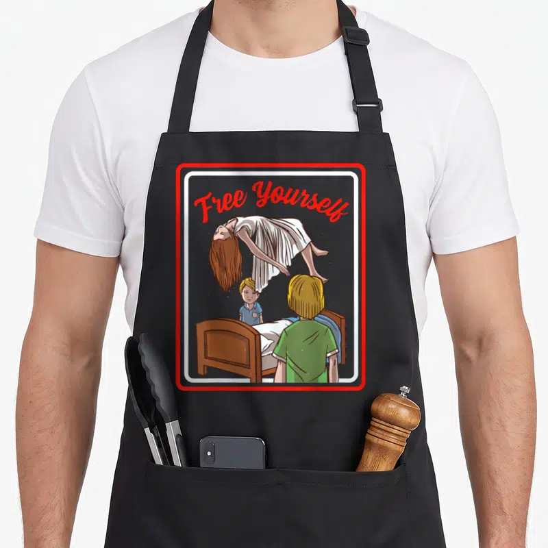 Tablier de cuisine noir avec illustration Free Yourself démon flottant au-dessus d'un lit et deux enfants.