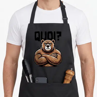 Tablier de cuisine noir avec ours brun musclé bras croisés et texte Quoi en lettres noires au-dessus.