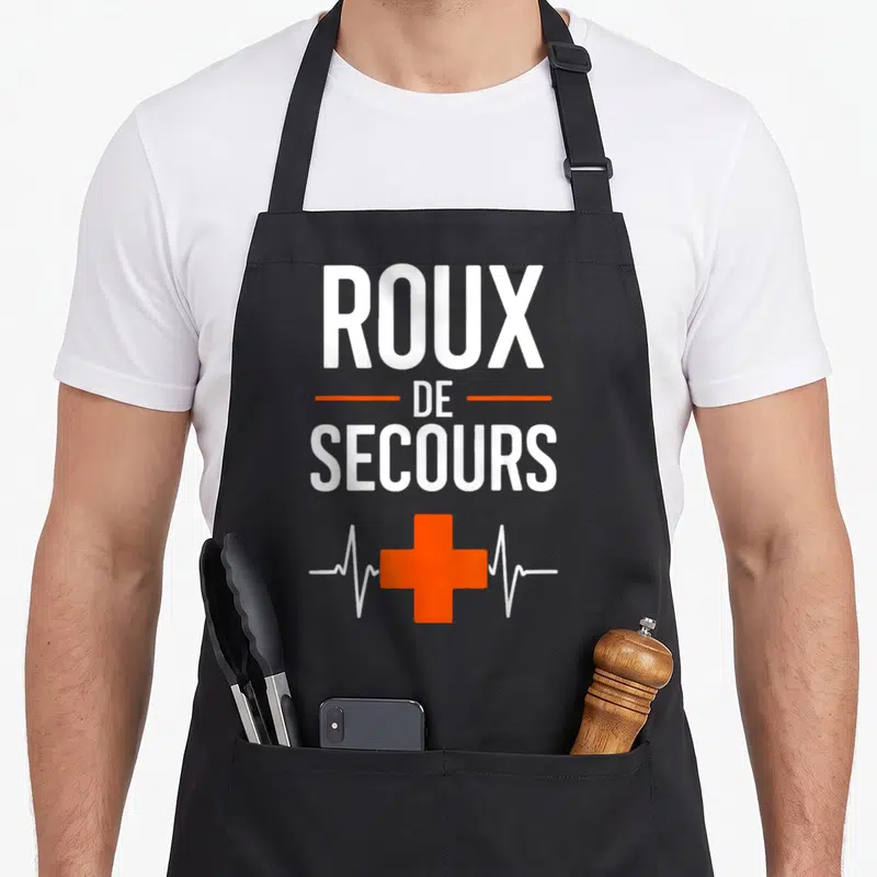 Tablier de cuisine noir avec inscription Roux de Secours et croix orange sur fond gris uni.