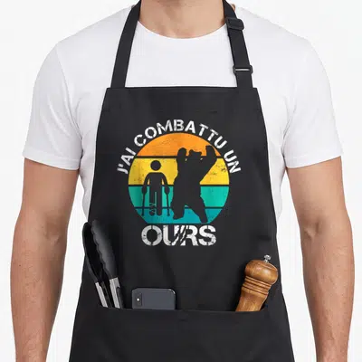 Tablier de cuisine noir avec illustration vintage d'un homme aux béquilles et d'un ours debout sur fond de soleil multicolore.