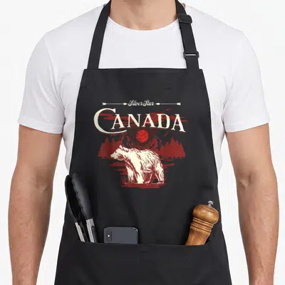 Tablier de cuisine noir avec illustration d'ours blanc, montagnes rouges et texte Canada en style vintage.