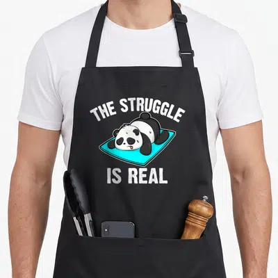 Tablier de cuisine noir avec un panda allongé sur un tapis bleu et le texte The Struggle Is Real imprimé en blanc.