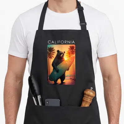 Tablier de cuisine noir avec un ours surfeur aux lunettes de soleil tenant une planche devant des palmiers au coucher du soleil.