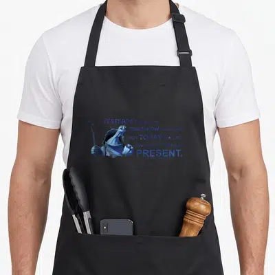 Tablier de cuisine noir avec portrait de tortue et texte en bleu évoquant le présent et l'histoire.