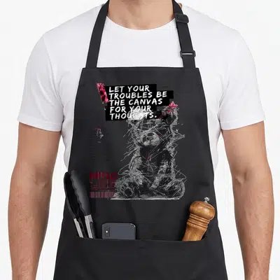 Tablier de cuisine noir avec illustration d'ours stylisé et texte blanc Let your troubles be the canvas for your thoughts en haut.