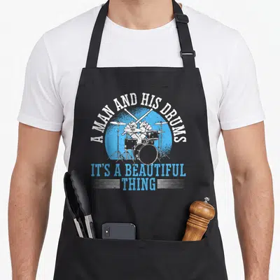 Tablier de cuisine noir avec illustration d'un batteur et texte A Man and His Drums It's une Chose Belle en bleu et blanc.
