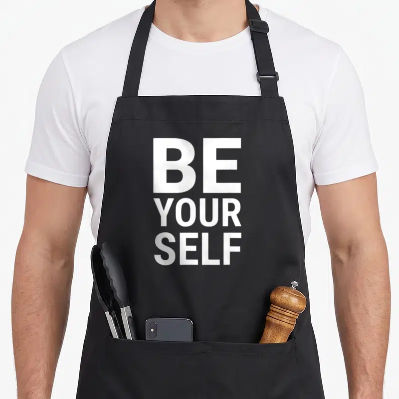 Tablier de cuisine noir avec le texte Be Yourself imprimé en grandes lettres blanches au centre.