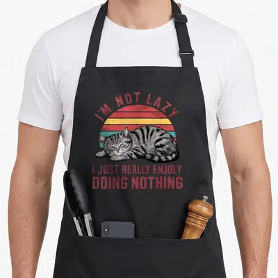 Tablier de cuisine noir avec illustration d'un chat gris dormant devant un soleil coloré et du texte en anglais.
