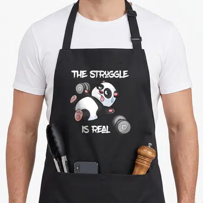 Tablier de cuisine noir avec un panda cartoon essoufflé tenant des haltères et le texte The Struggle Is Real.