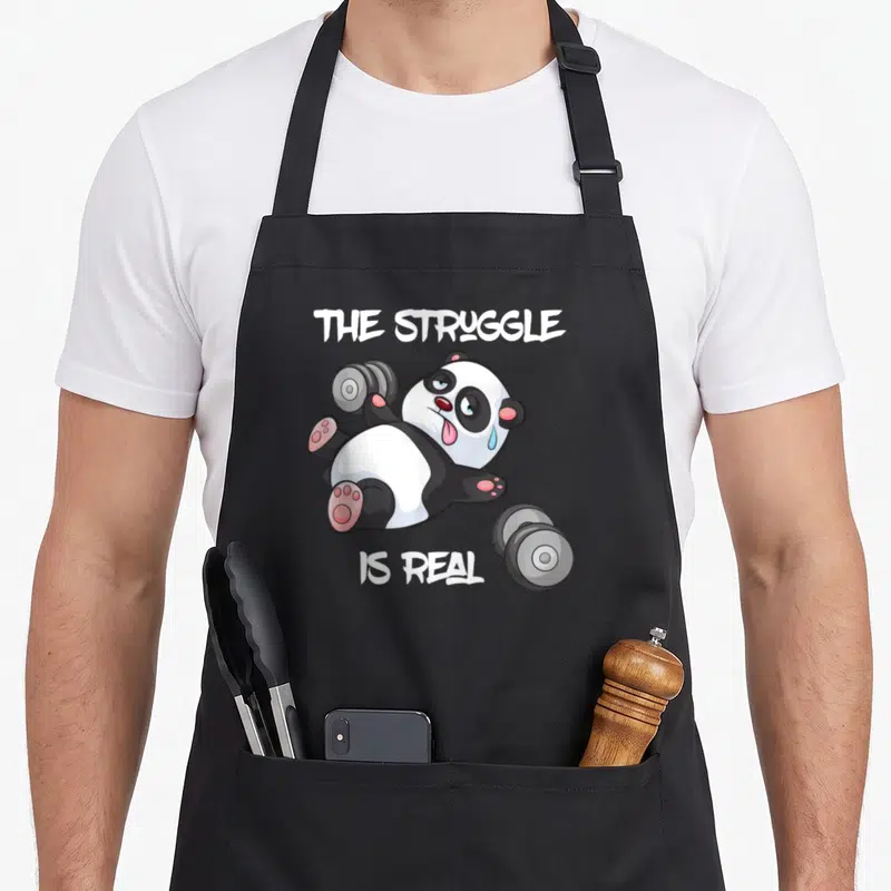 Tablier de cuisine noir avec un panda cartoon essoufflé tenant des haltères et le texte The Struggle Is Real.