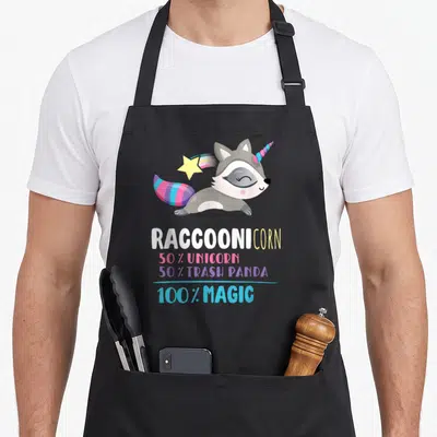 Tablier de cuisine noir avec un Raccoonicorn volant aux couleurs vives et des textes humoristiques en dessous.