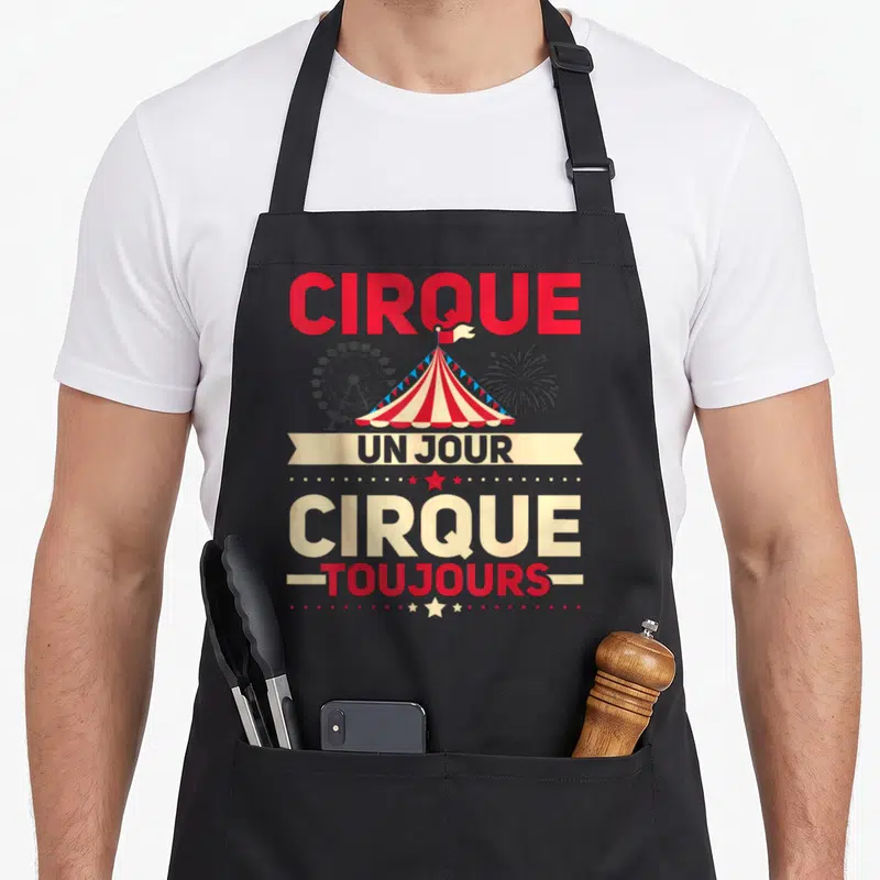 Tablier de cuisine noir avec une tente de cirque rouge et blanche, une grande roue et des feux d'artifice en arrière-plan.