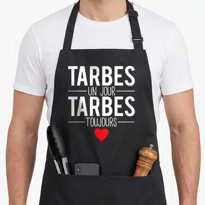 Tablier de cuisine noir avec l'inscription Tarbes un jour Tarbes toujours en blanc et un petit cœur rouge en dessous.