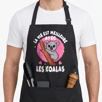 Tablier de cuisine noir avec un koala gris accroché à une branche sur fond rose et texte blanc.