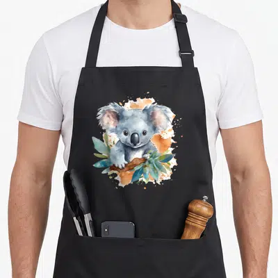 Tablier de cuisine noir avec une aquarelle représentant un koala gris sur une branche entourée de feuilles vertes.