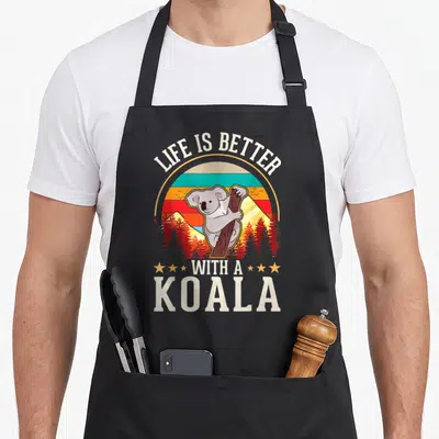 Tablier de cuisine noir avec illustration vintage d'un koala grimpant à un arbre devant un coucher de soleil rayé.