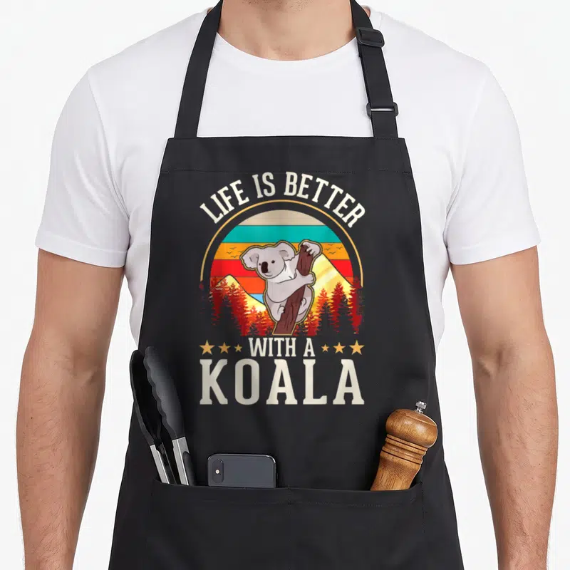 Tablier de cuisine noir avec illustration vintage d'un koala grimpant à un arbre devant un coucher de soleil rayé.