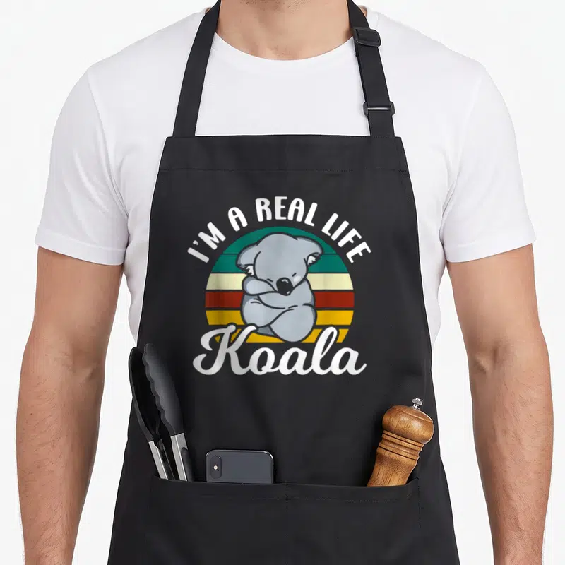 Tablier de cuisine noir avec illustration d'un koala gris aux bras croisés devant un soleil rayé multicolore et du texte blanc en anglais.
