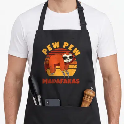 Tablier de cuisine noir avec un paresseux orange portant des lunettes de soleil et tenant deux revolvers sur fond de soleil couchant.