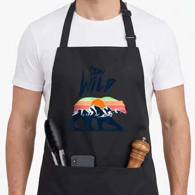 Tablier de cuisine noir avec un ours stylisé intégrant des montagnes et un soleil orange aux couleurs rétro.