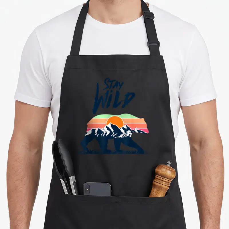 Tablier de cuisine noir avec un ours stylisé intégrant des montagnes et un soleil orange aux couleurs rétro.