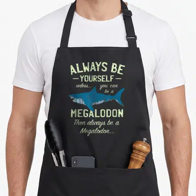 Tablier de cuisine noir avec l'illustration d'un Megalodon bleu et le texte Always be Yourself Unless you can be a Megalodon.