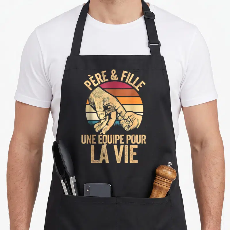 Tablier de cuisine noir avec illustration vintage d'une main d'adulte tenant celle d'un enfant devant un soleil couchant multicolore.