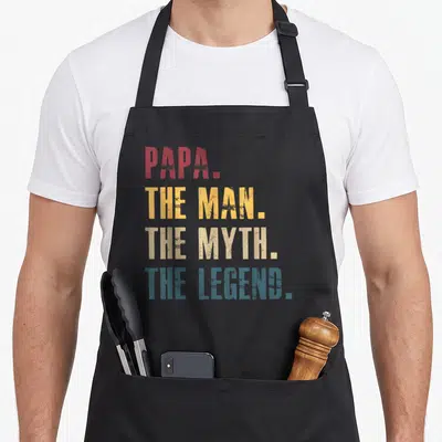 Tablier de cuisine noir avec linscription Papa The Man The Myth The Legend en lettres rouges jaunes et bleues.
