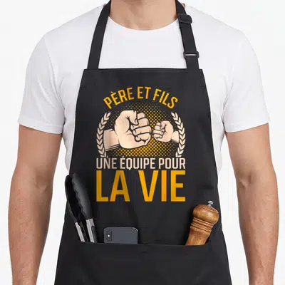 Tablier de cuisine noir avec deux poings se touchant et le texte Père et Fils une équipe pour la vie en orange et beige.