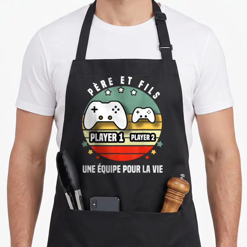 Tablier de cuisine noir avec deux manettes de jeu vidéo blanc et texte Père et Fils sur fond circulaire coloré.